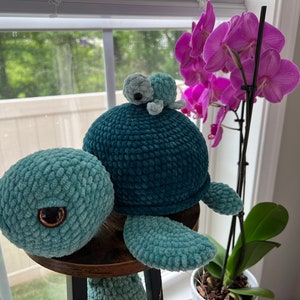 May include: A crocheted blue and green turtle with a smaller crocheted turtle on its back. The turtle is sitting on a wooden table in front of a window with a pink orchid plant.