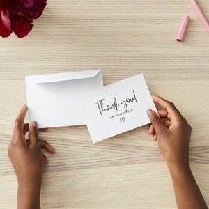 May include: A white thank you card with the text "Thank you! for your order" and a heart symbol is held by hands. An open white envelope is next to the card. A burgundy flower and a pink pen are in the background.