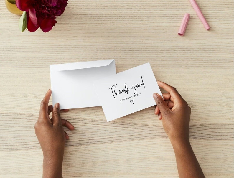 May include: A white thank you card with the text "Thank you! for your order" and a heart symbol is held by hands. An open white envelope is next to the card. A burgundy flower and a pink pen are in the background.