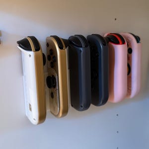 May include: A row of six game controllers in various colors, including white, gold, dark gray, and pink. The controllers are mounted on a white surface, with a black cable visible on the right side.