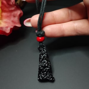 May include: A black obsidian pendant with a rough, textured surface hangs from a black cord with a red bead.