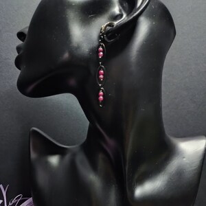 May include: A pair of dangle earrings with a black metal chain and pink beads. The earrings are on a black mannequin head.
