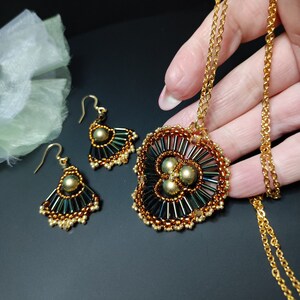 Può includere: Una collana con catena dorata con un ciondolo a forma di perla che presenta tre grandi perle verdi iridescenti circondate da perle più piccole dorate e nere. Sono inclusi anche orecchini abbinati con un design simile.