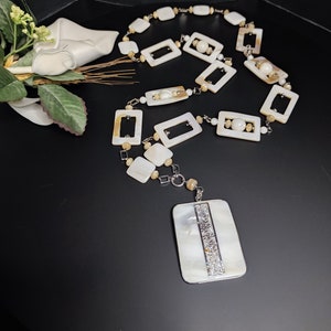 Può includere: Una lunga collana di conchiglie bianche e marroni con un ciondolo rettangolare. Il ciondolo ha una striscia argentata con strass.