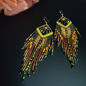 May include: A pair of handmade beaded earrings with a geometric design. The earrings feature a square top with a neon yellow border and gold accents. The fringe is a mix of brown, green, yellow, and gold beads, creating a layered effect.