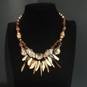 Peut inclure: Un collier multi-rangs avec des perles brunes, rouges et blanches. Le collier a un grand pendentif en coquillages blancs et bruns.