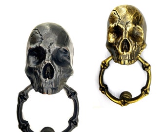 May include: Two skull-shaped door knockers. One is dark gray, the other is gold. Each has a ring-shaped handle. The skulls have detailed features, including eye sockets, a nose cavity, and a jawline. The door knockers are made of metal.