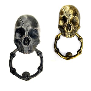 May include: Two skull-shaped door knockers. One is dark gray, the other is gold. Each has a ring-shaped handle. The skulls have detailed features, including eye sockets, a nose cavity, and a jawline. The door knockers are made of metal.