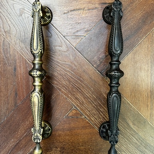 May include: Two ornate door handles. One is a brass color, the other is black. Both have a detailed, vintage design with a long, slender body and decorative circular backplates. The handles are set against a wooden background.