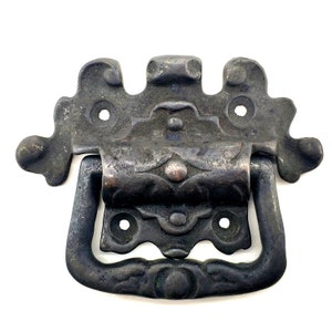 May include: Aged metal drawer pull with an ornate design and a dark finish. The pull has decorative flourishes and a curved handle. The hardware has a distressed appearance, suggesting a vintage or rustic style. Includes mounting holes for easy installation.