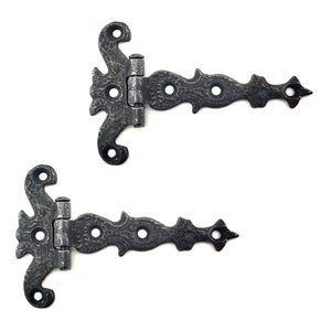 Puede incluir: Dos bisagras decorativas de estilo antiguo con un acabado oscuro y texturizado. Cada bisagra presenta un dise&ntilde;o ornamentado con una parte superior curvada, una bisagra central y un extremo puntiagudo. Las bisagras son de metal y est&aacute;n sobre un fondo blanco.