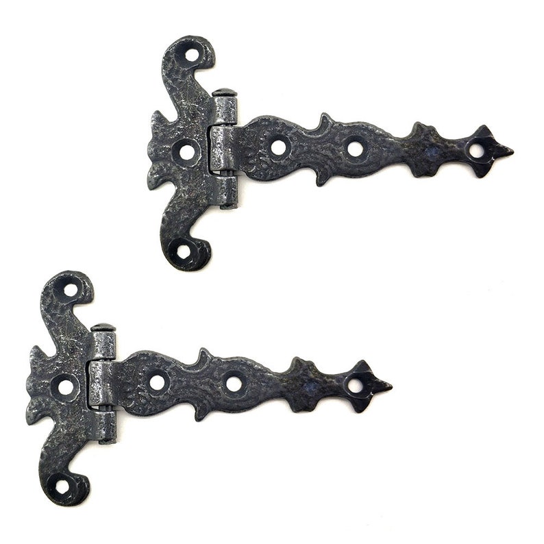 Puede incluir: Dos bisagras decorativas de estilo antiguo con un acabado oscuro y texturizado. Cada bisagra presenta un dise&ntilde;o ornamentado con una parte superior curvada, una bisagra central y un extremo puntiagudo. Las bisagras son de metal y est&aacute;n sobre un fondo blanco.