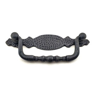 Op de afbeelding: Een donkergrijze metalen trekker met een gehamerde textuur. De trekker heeft een gebogen handvat met een decoratief midden en kogelvormige uiteinden. De hardware is ontworpen voor meubels en kasten.