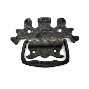 Può includere: Maniglia per cassetto in metallo antico con parte superiore decorativa e manico curvo. La parte superiore presenta un design ornato con tre sezioni e diversi fori di montaggio. La maniglia è di colore metallo scuro e invecchiato.