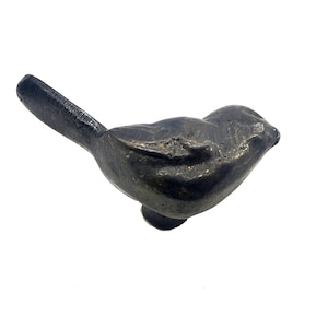 Op de afbeelding: Een klein, zwart metalen vogelbeeldje met een lange, dunne staart. De vogel bevindt zich in een rustpositie met zijn kop naar de zijkant gekanteld.