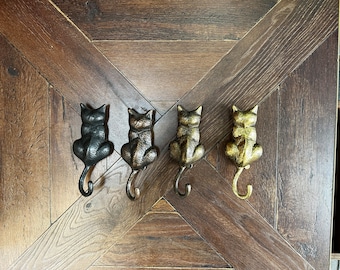 May include: Four black cast iron cat hooks in different finishes: black, bronze, silver, and gold. The hooks are shaped like cats sitting with their tails curled up.