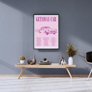 May include: A framed poster featuring the words "GETAWAY CAR" and a pink vintage car illustration. The poster is displayed on a wooden table with decorative items, including vases and a small plant. The background is a textured blue wall.