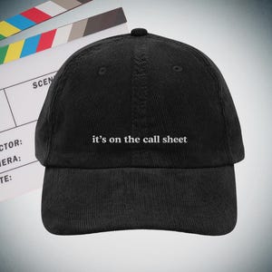 Może przedstawiać: Czarna sztruksowa czapka bejsbolowa z białym napisem "it's on the call sheet". W tle klaps filmowy. Czapka ma zakrzywiony daszek.