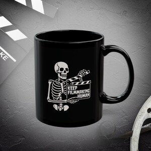 May include: Black ceramic mug featuring a white skeleton holding a film clapperboard. The clapperboard has the text "KEEP FILMMAKING HUMAN". A film clapperboard is in the background.