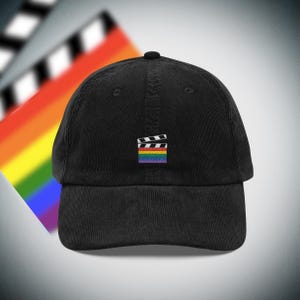 Könnte beinhalten: Schwarze Cord-Baseballkappe mit einem kleinen, gestickten Filmklappen-Logo in Regenbogenfarben. Die Kappe hat einen gebogenen Schirm und ein sechsteiliges Design. Der Hintergrund zeigt einen verschwommenen Regenbogenverlauf und eine Filmklappe.