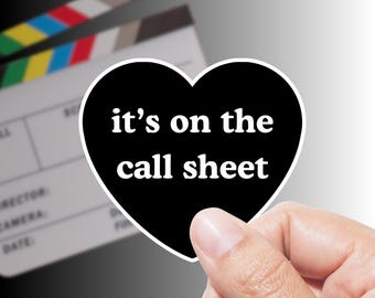 May include: A black heart-shaped sticker with the text "it's on the call sheet" in white. The sticker is held in a hand, with a film clapperboard in the background. The clapperboard has colorful stripes.