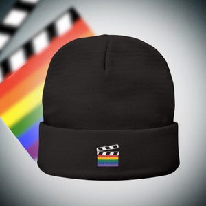 Op de afbeelding: Zwarte beanie met omgeslagen rand. De beanie heeft een geborduurde filmklapper met een regenboogvlag. De achtergrond bevat een vage filmklapper en een regenboogverloop.