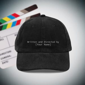 Op de afbeelding: Zwarte corduroy baseball cap met de tekst "Written and Directed by [Your Name]" in het wit. Een filmklapper is op de achtergrond te zien. De cap is een klassieke, verstelbare stijl.