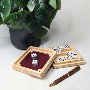 May include: Wooden dice game with a burgundy felt interior, featuring white dice with black dots. A wooden pen with gold accents rests nearby. A large, dark gray pot with a green plant is in the background.