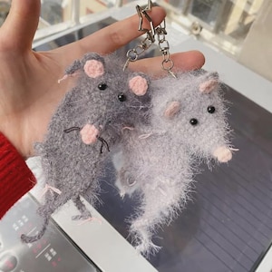 May include: Two fuzzy, crocheted rat keychains attached to a star-shaped metal ring. One rat is dark gray and the other is light gray, both featuring small pink crocheted ears, pink noses, black bead eyes, and thin, textured yarn fibers.