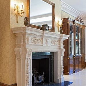 May include: A white marble fireplace with ornate carvings, a dark green hearth, and a decorative metal grate. A large framed mirror hangs above the mantel, with sconces and a clock. A wooden cabinet is to the right.