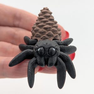 May include: A 3D-printed figurine of a spider with a pinecone-shaped head. The spider has a black body and legs, with a brown pinecone head. The eyes are small and white. The figurine is held in a hand.