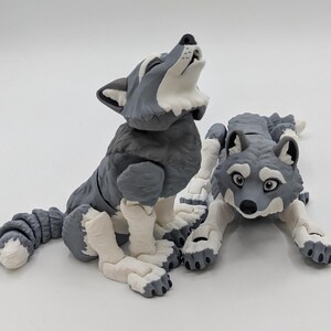 May include: Two gray and white wolf figurines. One wolf is sitting up and howling, while the other is lying down. The figurines have detailed fur textures and are made of a smooth material. The wolves are a charming decorative item.