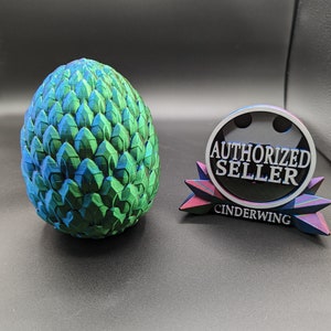 May include: A green and blue dragon egg replica with a textured surface. The egg is next to a black and silver circular badge with the text "AUTHORIZED SELLER" and "CINDERWING" printed on it.