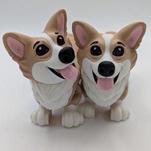 May include: Two smiling corgi figurines. The corgis have tan and white fur, black eyes, and pink tongues and ears. The figurines are standing side-by-side, with their tongues sticking out. The corgis have white paws and are on a white surface.