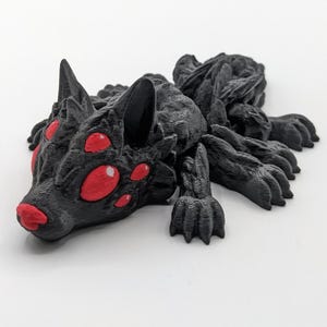 May include: A black 3D printed figurine of a wolf-like creature with multiple red eyes. The creature has a textured surface and appears to be crawling.