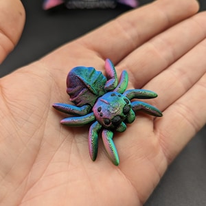 May include: A small, iridescent green and purple spider figurine with black eyes. The spider is made of a textured material and has a detailed design.