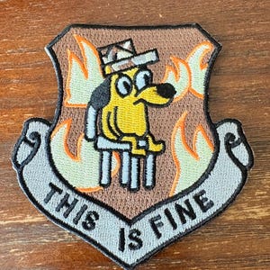 May include: An embroidered shield-shaped patch featuring a yellow dog wearing a hat, sitting on a chair amidst orange and yellow flames. Below the dog, a grey banner displays the black text THIS IS FINE in capital letters.