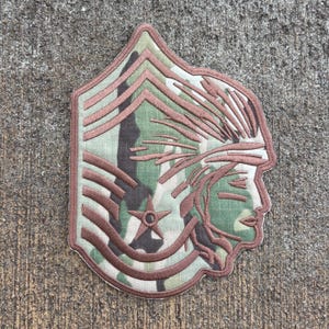 Puede incluir: Un parche bordado de camuflaje marrón y verde con un perfil de un nativo americano que lleva un tocado de plumas. El parche tiene una estrella de cinco puntas en el centro y un patrón de chevron alrededor de los bordes.