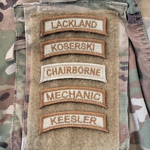 May include: A camouflage fabric patch with five brown embroidered patches that read "Lackland", "Koserski", "Chairborne", "Mechanic", and "Keesler".