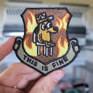 May include: A brown, green, and yellow embroidered patch with a cartoon dog wearing a hat and sitting in flames. The patch has the text "This is fine" on it.