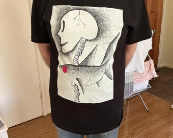May include: Black t-shirt with a white graphic print of a skull with a cracked head and a fish with a red mouth. The fish is attached to the skull by a chain.