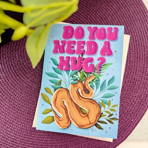 May include: A greeting card with a light blue background featuring a snake with a brown and orange pattern. The snake is surrounded by green leaves and the text "Do you need a hug?" in pink letters.