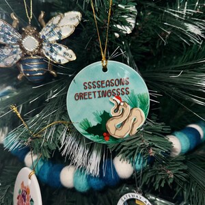 Op de afbeelding: Een ronde, groene kerstbal met een witte slang die een rode kerstmanmuts draagt. De bal heeft de tekst "SSSSSEASONS GREETINGSSSS" erop.