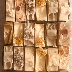 Könnte beinhalten: Eine Sammlung rechteckiger handgemachter Seifen in verschiedenen Beige-, Creme- und Brauntönen, angeordnet in Reihen. Einige Seifen sind mit getrockneten Blumen oder kleinen Rosenknospen verziert. Die Seifen werden in einzelnen braunen Papierbehältern präsentiert.