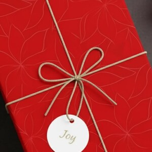 May include: A wrapped gift box with red floral patterned paper, tied with twine and a tag that says "Joy". The box is surrounded by Christmas ornaments and greenery.