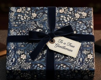May include: A wrapped gift box with a floral pattern in navy blue and white, tied with a navy blue velvet ribbon and bow. A tag reads "To a True Gentleman". Gold cufflinks and a leather-bound notebook are also visible.