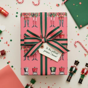 May include: A wrapped gift with pink wrapping paper featuring a nutcracker soldier pattern. The gift is tied with a green and pink striped ribbon and a tag that reads, "To the jolliest person I know!"