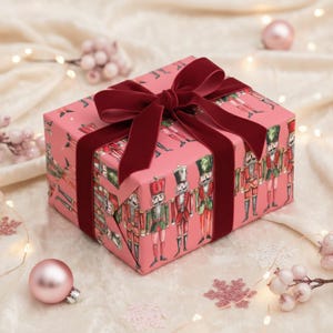 May include: A wrapped gift box with pink wrapping paper featuring a repeating pattern of toy soldiers. The box is tied with a burgundy velvet ribbon. Decorative Christmas ornaments and lights surround the gift.