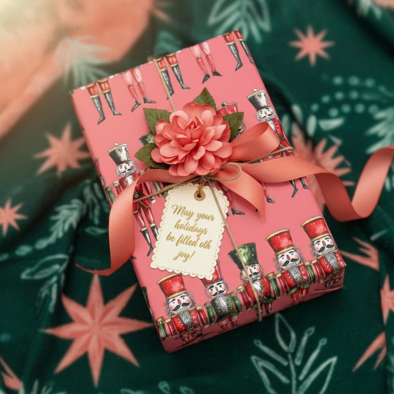 May include: A wrapped gift with pink wrapping paper featuring a nutcracker soldier pattern. The gift is adorned with a peach-colored ribbon, a paper flower, and a tag that reads "May your holidays be filled with joy!"