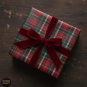 Può includere: Un regalo avvolto in carta a quadri rossi e verdi, legato con un ampio nastro e fiocco di velluto rosso scuro. Il regalo è su una superficie di legno scuro. Il logo Studio Select è visibile in basso a sinistra.